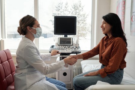 Side view of female clinician and patient shaking hands in medical office while sitting in front of one another against ultrasound machine