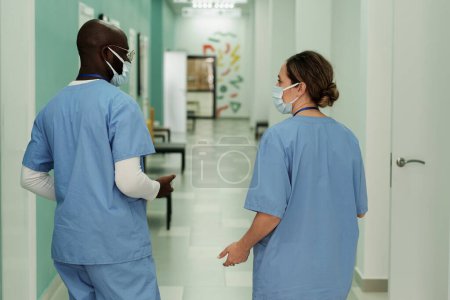 Téléchargez les photos : Rear view of two young interracial clinicians in blue uniform and protective masks having discussion in hospital corridor - en image libre de droit