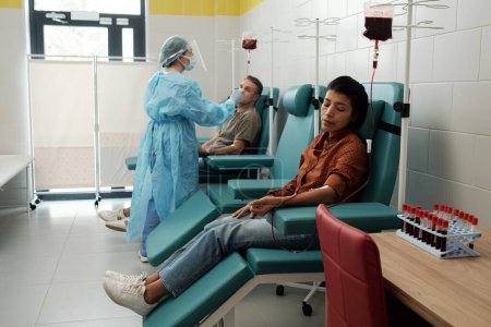 Téléchargez les photos : Young Hispanic female donor giving her blood for sick patients of hospital while sitting in armchair in large ward - en image libre de droit