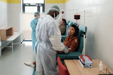 Foto de Young Hispanic female donor looking at African American clinician in protective coveralls, gloves, face screen and headband - Imagen libre de derechos