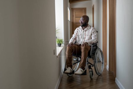 Foto de Young African American man with disability looking through window while sitting on wheelchair in corridor of apartment - Imagen libre de derechos