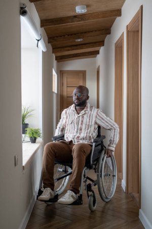 Téléchargez les photos : African American man with disability sitting on wheelchair in long corridor of large modern apartment and looking at camera - en image libre de droit