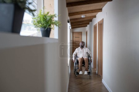 Téléchargez les photos : Young man with disability in casualwear moving along corridor of modern apartment while sitting on wheelchair - en image libre de droit