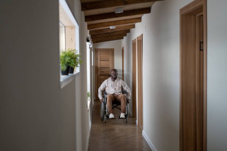Foto de Young man of African ethnicity with disability moving along corridor of contemporary apartment while sitting in wheelchair - Imagen libre de derechos