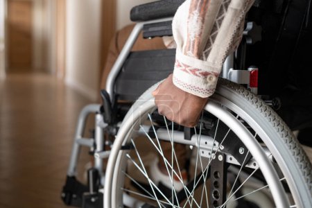 Téléchargez les photos : Close-up of man with disability rotating wheels of wheelchair while moving forwards along corridor of his apartment - en image libre de droit