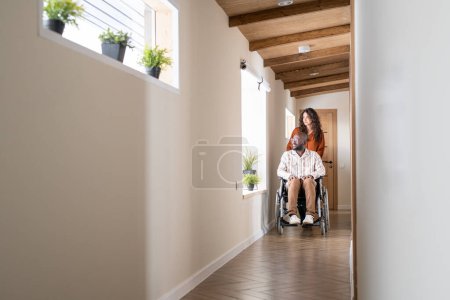 Téléchargez les photos : Young female caregiver pushing wheelchair with African American man with disability while both looking through window - en image libre de droit