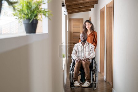Téléchargez les photos : Young contemporary interracial couple standing in corridor of apartment while female caregiver standing in front of man in wheelchair - en image libre de droit