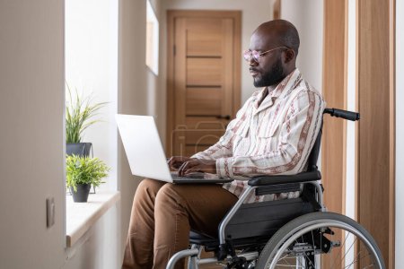 Foto de Young serious black man with disability concentrating on network while sitting in wheelchair in corridor of apartment - Imagen libre de derechos