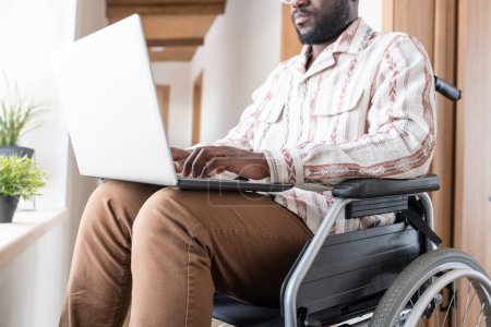 Téléchargez les photos : Close-up of young man with disability working in the net in home office while sitting in wheelchair in corridor of modern apartment - en image libre de droit