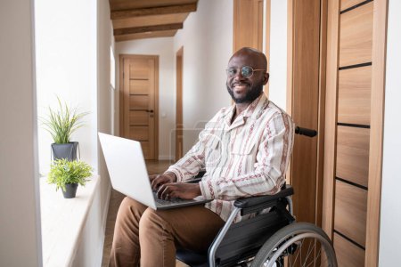 Téléchargez les photos : Young successful African American businessman with laptop on his knees looking at camera while sitting on wheelchair and networking - en image libre de droit
