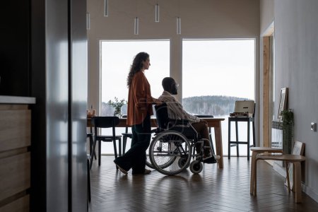 Photo for Side view of young female caregiver pushing wheelchair with African American man while moving along spacious living room - Royalty Free Image