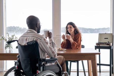 Téléchargez les photos : Happy young female with mug sitting by kitchen table and looking at black man with disability during conversation by breakfast - en image libre de droit