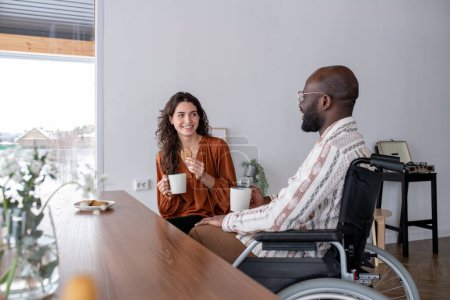 Téléchargez les photos : Cheerful female caregiver with cup of tea and cookie looking at black man with disability in wheelchair while talking to him - en image libre de droit