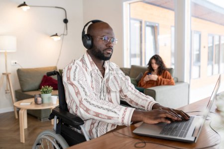 Foto de Serious African American businessman in headphones typing by table in living room while working with online data against young woman - Imagen libre de derechos
