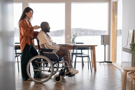 Téléchargez les photos : Young woman with electric trimmer cutting hair on head of man with disability sitting in wheelchair in spacious living room - en image libre de droit