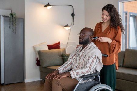 Foto de Young attractive female hairdresser or caregiver cutting hair of man in wheelchair while standing behind him in living room - Imagen libre de derechos