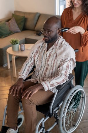 Foto de Young smiling female caregiver in casualwear cutting hair of black man in wheelchair with electric clippers while standing behind - Imagen libre de derechos