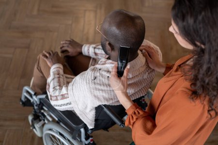 Téléchargez les photos : Above angle of black man with disability sitting in wheelchair while having his hair cut by female caregiver or hairdresser - en image libre de droit