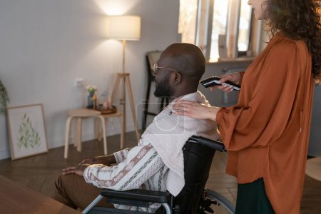 Téléchargez les photos : Young African American man with disability having his hair cut by electric trimmer held by female caregiver standing behind him - en image libre de droit