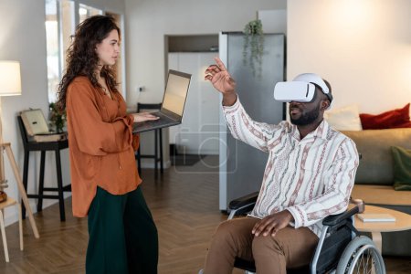 Téléchargez les photos : Young woman with laptop standing in front of man with disability sitting in wheelchair with vr headset and pointing at virtual screen - en image libre de droit