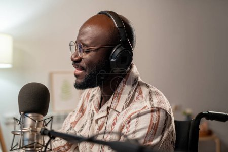 Foto de Young bald African American man in eyeglasses and headphones singing in microphone while recording his songs in studio - Imagen libre de derechos