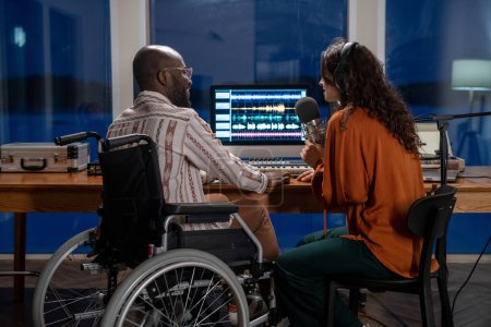 Photo for Young African American man in wheelchair looking at female in headphones singing in microphone while recording her songs - Royalty Free Image