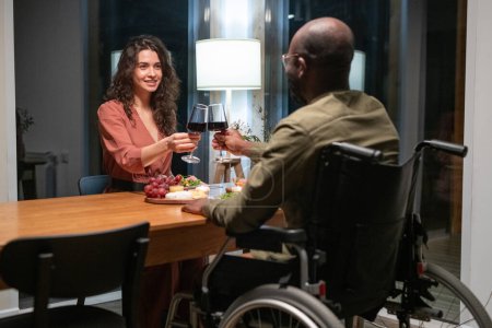 Téléchargez les photos : Happy young interracial couple sitting by festive table and toasting with red wine during romantic dinner at home or restaurant - en image libre de droit