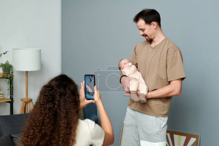 Foto de Happy young man looking at baby son on hands while standing in front of his wife taking photo of them on smartphone - Imagen libre de derechos