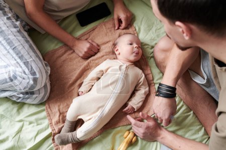 Foto de Above view of cute baby boy in white romper grimacing while relaxing on bed between his mother and father playing with him - Imagen libre de derechos