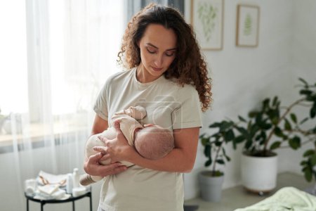 Foto de Contemporary young mother in t-shirt holding newborn baby by her chest while lulling him in bedroom in the middle of day - Imagen libre de derechos