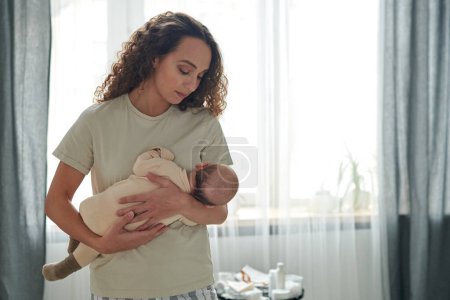 Foto de Young serene woman with her baby son by her chest lulling him while standing in bedroom against white and grey curtains - Imagen libre de derechos