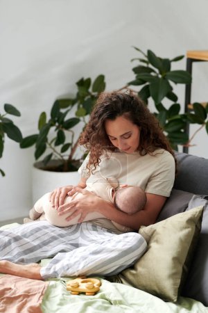 Téléchargez les photos : Young mother in homewear relaxing by cushions on double bed in bedroom and putting her cute baby son to sleep - en image libre de droit