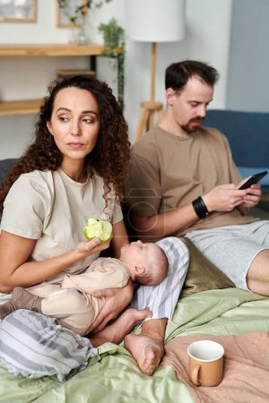 Young serene female with sleeping baby eating fresh green apple for breakfast while sitting on double bed against her husband