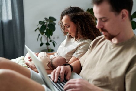 Photo for Young serene woman with newborn baby son on hands sitting on bed by her husband typing on laptop keyboard while networking - Royalty Free Image