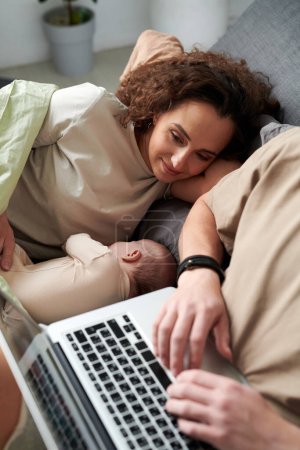 Photo for Young serene woman looking at cute baby son lying next to her during relaxation on bed in front of man typing on laptop keyboard - Royalty Free Image