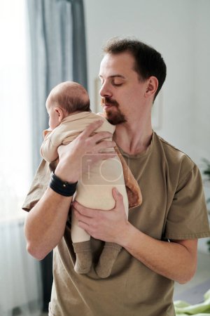 Photo for Young bearded man in t-shirt looking at baby son in white bodysuit on his shoulder while lulling him against curtains on window - Royalty Free Image