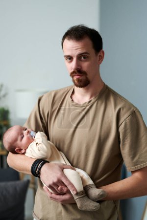 Foto de Young man in casual t-shirt holding baby boy in romper suit while standing in front of camera and lulling infant - Imagen libre de derechos