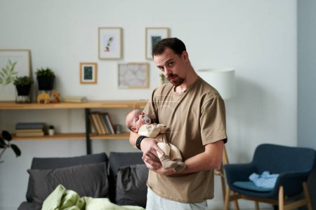 Téléchargez les photos : Contemporary young man lulling baby son while standing in front of camera against shelves, pictures in frames, armchair and bed - en image libre de droit