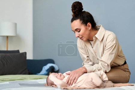 Foto de Young contemporary female in smart casualwear bending over her baby son lying on double bed while going to get him dressed - Imagen libre de derechos