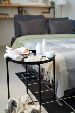 Téléchargez les photos : Set of white plastic bottles with body lotions and creams, nappies and cotton pads in glass container on small table against double bed - en image libre de droit