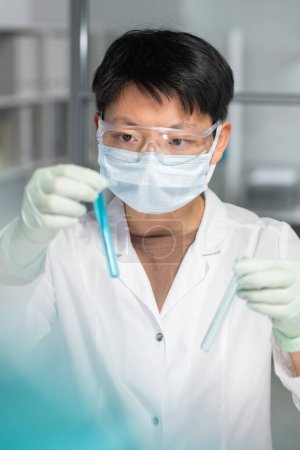 Photo for Young female scientist of Asian ethnicity holding flask with blue liquid in hand and looking at it during scientific research - Royalty Free Image
