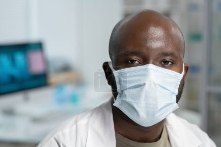 Photo for Head of young black man in whitecoat protective mask looking at camera while sitting by workplace in modern scientific laboratory - Royalty Free Image