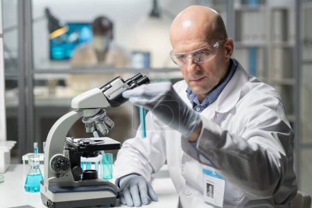 Foto de Contemporary mature male clinician in gloves, whitecoat and protective eyewear looking at blue liquid in flask while sitting in laboratory - Imagen libre de derechos