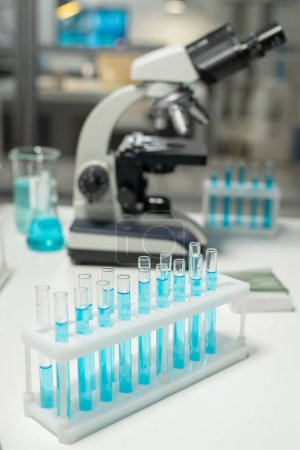 Photo for Group of flasks with samples of blue liquid standing on workplace of scientist against microscope and other glass equipment - Royalty Free Image