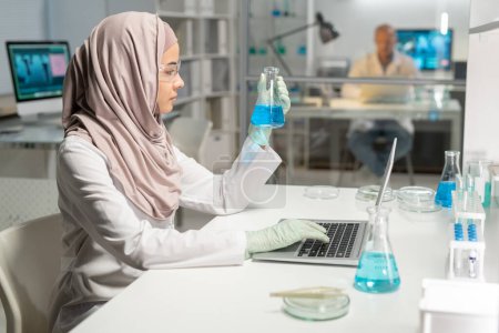 Foto de Side view of young Muslim female scientist in hijab looking at test tube with blue liquid in her gloved hand during scientific research - Imagen libre de derechos
