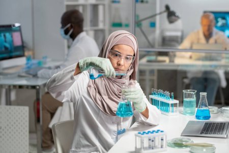 Photo for Young serious Muslim female researcher in hijab, gloves and protective eyewear pouring liquid substance into flask with blue fluid - Royalty Free Image