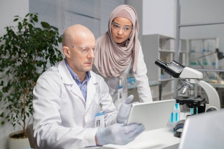 Photo for Two young contemporary virologists discussing online data in laboratory whilemature male scientist pointing at tablet screen - Royalty Free Image