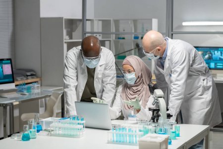 Photo for Young Muslim female researcher in hijab, whitecoat, protective gloves, mask and eyeglasses making presentation to male colleagues - Royalty Free Image