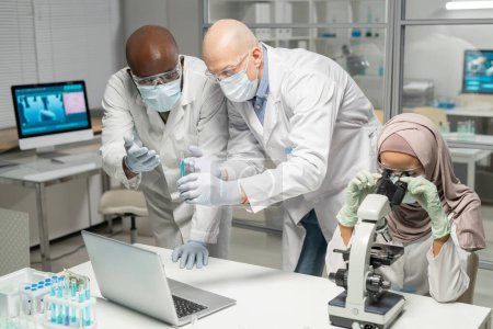Photo for Interracial male scientists discussing blue liquid in flask next to young Muslim female virologist studying new virus in microscope - Royalty Free Image
