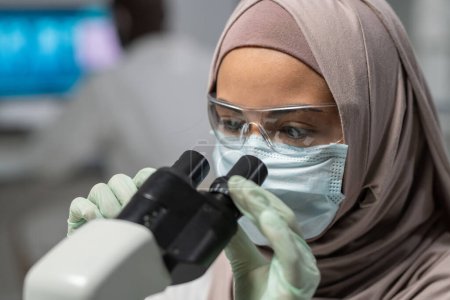 Photo for Close-up of young Muslim female researcher looking in microscope during scientific investigation or discovery of new virus - Royalty Free Image